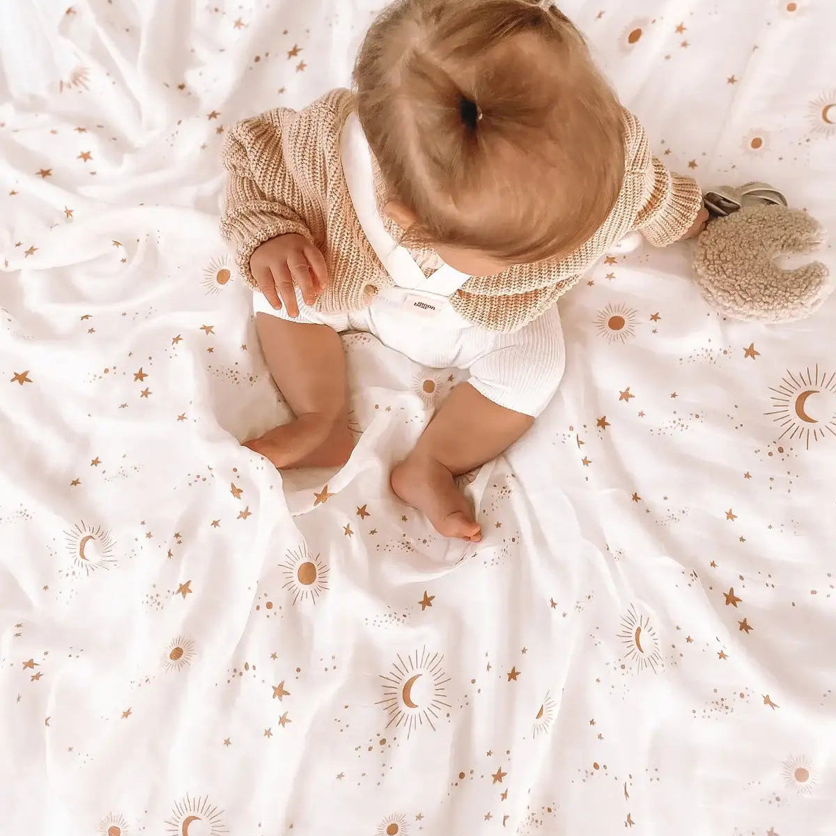 Baby sitting on a large white Constellation muslin swaddle
wearing a knit cardigan, top down view
