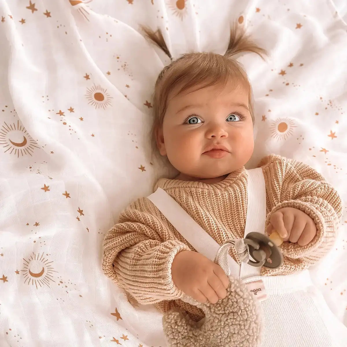 Baby lying on a Constellation print organic muslin swaddle holding a pacifier clip, close up